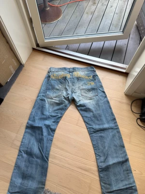 Ljusblå replay med brodyr baktill - Säljer ett par ljusblå jeans från Replay med gul brodyrtext på bakfickorna. Jeansen har raka ben, slitna detaljer och klassisk femficksdesign. Materialet är denim och de har en avslappnad streetwear-look.