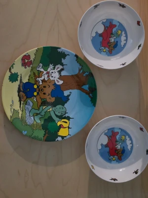Bamse tallrik och skålar set - Säljer ett set med en tallrik och två skålar med färgglada Bamse-motiv. Tallriken har en tecknad skogsscen med Bamse och hans vänner, medan skålarna har motiv av Bamse i ett flygplan. Perfekt för barn och samlare av Bamse-produkter.