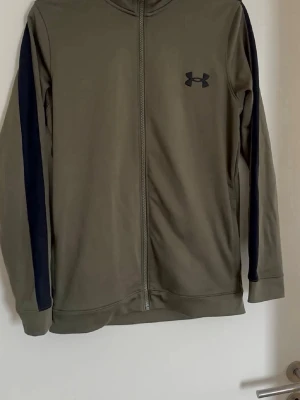 Grönt träningsset från Under Armour - Snyggt träningsset från Under Armour i olivgrönt med svarta detaljer längs ärmar och ben. Setet består av en ziptröja med hög krage och matchande byxor med dragsko i midjan. Perfekt för gymmet eller löprundan.