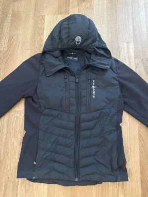 Sail Racing Jacka - Snygg svart vindjacka från Sail Racing med vadderad front och rygg, två bröstfickor med dragkedja och stor huva. Jackan har slimmad passform och är tillverkad i ett lätt och vindtätt material. Perfekt för blåsiga dagar och outdoor-aktiviteter. Passar både Höst och Vinter!