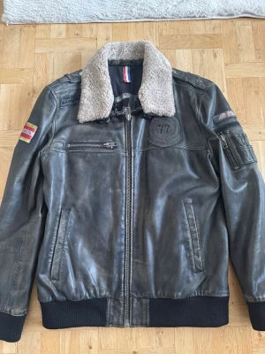 Svart skinnjacka med fårkrage - Snygg svart skinnjacka med mjuk fårkrage och detaljerade patchar på bröst och ärm. Jackan har dragkedja framtill, flera fickor och ribbade muddar. Materialet är äkta läder och kragen är i fuskpäls. Perfekt för dig som gillar en cool och tidlös stil.