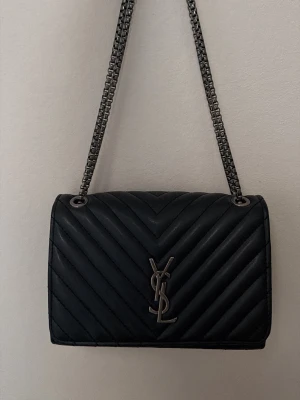 Svart YSL handväska i läder - Snygg svart handväska från YSL i quiltat läder med silverfärgad kedjerem och YSL-logga framtill. Väskan har en stilren, rektangulär form och passar perfekt till en trendig look. Kedjan kan bäras både på axeln och crossbody.