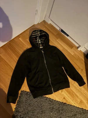 Svart hoodie med dragkedja - Svart hoodie med hel dragkedja och huva. Insidan av huvan har ett rutigt mönster i svart, vitt och beige. Hoodien har två stora fickor framtill och ribbade muddar vid ärmslut och nederkant. Perfekt för en avslappnad stil.