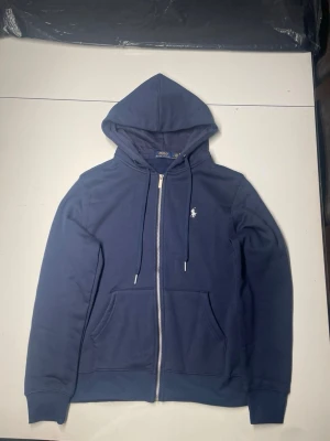 Blå zip-hoodie  - Mörkblå hoodie med dragkedja. Klassisk design med huva, dragsnören och två fickor framtill. Liten vit logga broderad på bröstet. Perfekt för en avslappnad och stilren look.