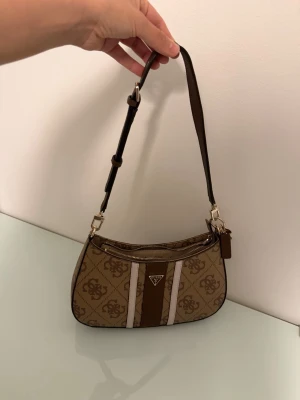 Brun Guess axelremsväska  - Snygg brun handväska från Guess med justerbar axelrem och klassiskt logomönster. Väskan har två vita ränder framtill, guldfärgade detaljer och dragkedja både fram och bak. Perfekt storlek för det viktigaste och en trendig accessoar till din outfit. Köpte från Zalando i maj. Har använt sparsamt är som ny. Pris kan diskuteras vid snabb affär