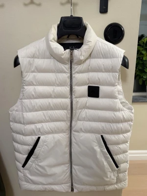 Vit dunväst från moose knuckles - Snygg vit dunväst från Moose knuckles med quiltad design och svarta dragkedjor på fickorna. Västen har hög krage och en diskret svart patch på bröstet. Perfekt för lager-på-lager och ger en clean look till din outfit.