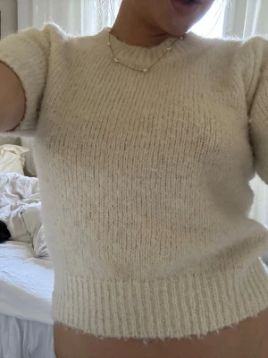 Krämvit stickad t-shirt - Kortärmad stickad tröja i krämvit färg med rund hals och ribbade kanter. Tröjan har en mjuk, fluffig struktur och är något croppad i modellen. Perfekt för dig som gillar en enkel men trendig look.