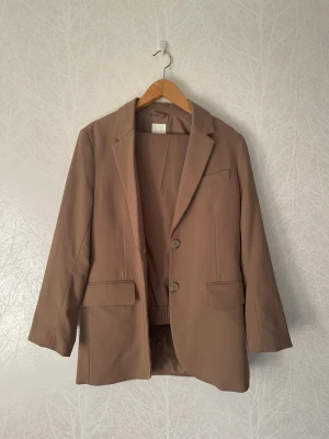 Ljusbrunt H&M Kavajset - Ljusbrunt oversized kavajset från H&M som använts ett par gånger men är i mycket bra skick. En trasig söm fanns i fodret, men den har sytts ihop, och den syns knappt. Byxorna har dessutom sytts lite kortare så att de passar någon som är 160 cm.  Kavajen är i storlek XS och byxorna i storlek S.   Längd Kavaj: ca 75 cm. Axelbredd Kavaj: ca 42 cm.  Längd Byxor: ca 95 cm.   Säljs tillsammans.