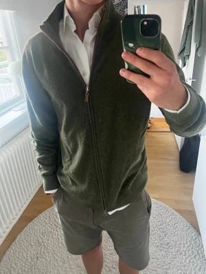Olivgrön ziptröja från Less Worries - Olivgrön stickad ziptröja från Less Worries med hög krage och hel dragkedja. Tröjan har ribbade muddar och en diskret röd detalj längs dragkedjan. Perfekt att bära över en skjorta för en stilren look. Denna är riktigt snygg. Köpte för 800 tror jag, knappt använd