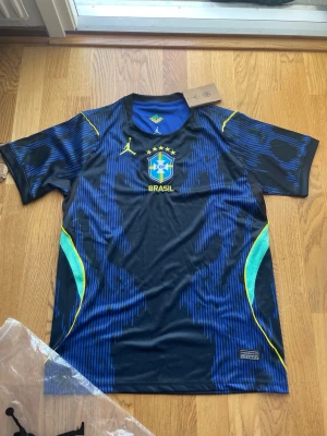 Blå Brasilien matchtröja Jordan - Snygg blå och svart Brasilien matchtröja från Jordan med gula detaljer och CBF-emblem på bröstet. Tröjan har korta ärmar, rund hals och är tillverkad i ett lätt, ventilerande material som passar perfekt till fotboll eller träning.