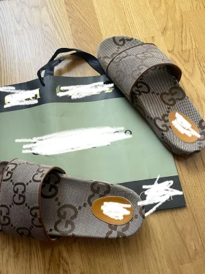 Bruna sandaler med GG-mönster - Säljer ett par bruna sandaler med klassiskt GG-mönster i beige och mörkbrunt. Sandalerna har bred rem över foten och en oval detalj i brunt och beige framtill. Sulan är räfflad undertill för bättre grepp. Perfekta för sommaren och riktigt snygga till streetwear.