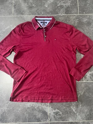 Vinröd långärmad pikéskjorta - Snygg och stilren långärmad pikétröja från Tommy Hilfiger i en djup vinröd nyans. Tröjan har en slim fit-passform, snygga randdetaljer på krage och knäppning samt den klassiska småbroderade loggan på bröstet. 