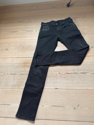 Svarta Replay Anbass jeans - Säljer ett par svarta Replay Anbass jeans i slim fit-modell. Jeansen har klassisk femficksdesign, knappgylf och är tillverkade i stretchigt material för extra komfort. Perfekta för dig som gillar en clean och stilren look.