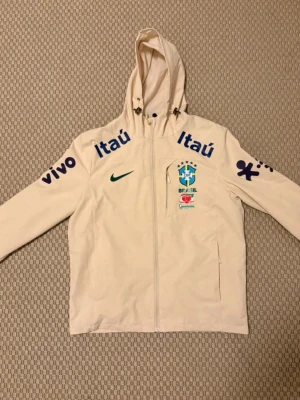 Vit Brasil Nike vindjacka med huva - Snygg vit vindjacka från Nike med Brasil-logga och sponsormärken som Itaú och Vivo. Jackan har huva med dragsko, hel dragkedja framtill och två sidofickor. Perfekt för dig som gillar sportig stil och vill sticka ut med landslagskänsla.