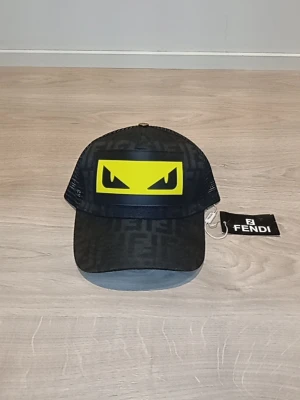 Svart keps med gult monstertryck Fendi - Snygg svart keps från Fendi med ikoniskt gult monsterögon-tryck framtill. Kepsen har mesh på sidorna och bak, samt justerbar rem. Materialet är en mix av textil och mesh, med diskret Fendi-mönster över hela kepsen. Perfekt för dig som vill sticka ut.