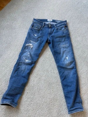 Blå slitna replay jeans - Säljer ett par blå replay jeans med slitningar och distressed detaljer på benen. Jeansen har klassisk femficksdesign och normal midja. Perfekta för en avslappnad och trendig look. Det är 29 waist och 30 length och passar också om man skulle ha större midja, har du en fråga var då inte rädd att ställa den