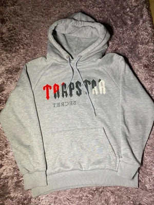 Grå hoodie med TRAPSTAR-tryck - Snygg grå hoodie med stor huva och magficka. Framtill står det 'TRAPSTAR' i svart, vitt och rött tryck samt texten 'IT'S A SECRET' under. Tillverkad i mjukt bomullsmaterial och har ribbade muddar vid ärmar och nederkant. Perfekt för en avslappnad streetwear-look.