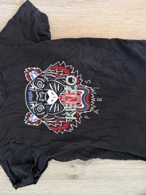 Svart Kenzo t-shirt med tigertryck - Svart t-shirt från Kenzo med ikoniskt tigertryck i vitt, rött och blått på bröstet. Klassisk passform och rund hals. Perfekt för dig som vill sticka ut med en statement tee.