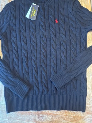 Mörkblå kabelstickad tröja Polo Ralph Lauren - Kabelstickad mörkblå tröja från Polo Ralph Lauren med rund halsringning och ribbade muddar. Tröjan har det klassiska röda Polo-logot på bröstet och är tillverkad i mjukt material. Perfekt för dig som gillar stilrena och tidlösa plagg.