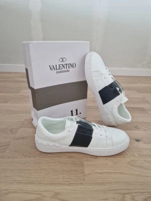 Valentino open  - Stilrena vita sneakers från Valentino Garavani med bred svart rem över sidan och nitar vid hälen. Skorna har en klassisk låg siluett, vita snören och en tjock vit sula. Perfekta för dig som vill ha en lyxig och modern look. Aldrig använda. Box ingår ej!