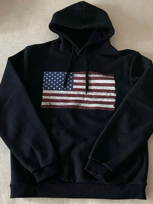 Svart hoodie med amerikansk flagga - Säljer en svart hoodie med huva och dragsnören. På bröstet finns ett tryck av den amerikanska flaggan i rött, vitt och blått med en lite sliten look. Hoodien har ribbade muddar vid ärmar och nederkant. Perfekt för dig som gillar streetwear och vill sticka ut. Storlek M men kan passa S.