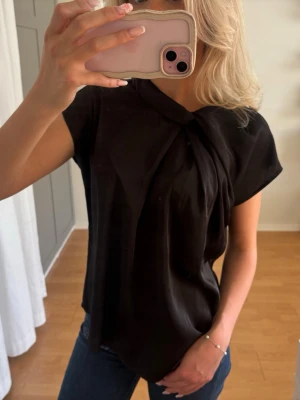 Satintopp Neo Noir  - Säljer en elegant svart blus med knytning framtill. Blusen har korta ärmar och ett fint fallande tyg som ger en stilren och feminin look. Perfekt till både vardag och fest! Använd fåtal gånger och i mycket fint skick.
