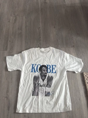 Vit t-shirt med Kobe Bryant-tryck - Vit t-shirt med stort tryck av basketspelaren Kobe Bryant och texten 'KOBE' i blått. Klassisk passform och rund halsringning. Perfekt för dig som gillar sport och streetwear.