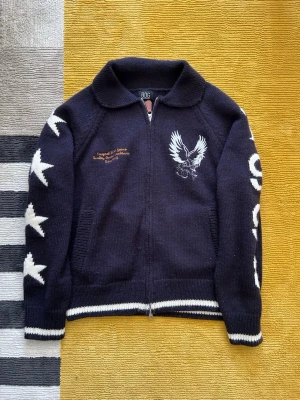 Vintage tröja/kofta - Mörkblå stickad ziptröja från BDG med vita stjärnor på ärmarna, broderad örn på bröstet och textdetaljer både fram och bak. Tröjan har ribbade muddar och krage samt vita ränder vid ärmslut och nederkant. Perfekt för dig som gillar streetwear och unika detaljer. Använt 2 gånger och är från urban outfits, ny pris 1200