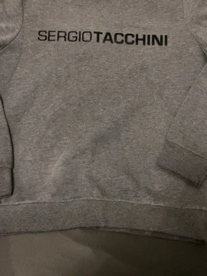 Grå sweatshirt från Sergio Tacchini - Snygg grå sweatshirt från Sergio Tacchini med svart logotyptryck över bröstet och en liten logga på ärmen. Tröjan har rund halsringning, ribbade muddar och är gjord i mjukt bomullsmaterial. Perfekt för en chill och sportig look.