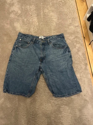 Blå jeansshorts från Grant - Säljer ett par klassiska blå jeansshorts från Grant. De har fem fickor, normal passform och är tillverkade i slitstark bomull. Perfekta för sommaren och har en tidlös look med knappgylf och bälteshällor.