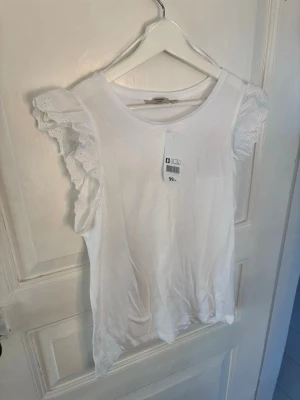 Vit ärmlös topp med volangdetaljer - Säljer en söt vit topp med volangdetaljer på axlarna. Toppen är ärmlös och har broderade spetsvolanger som ger en romantisk vibe. Perfekt att styla med jeans eller kjol för en fräsch look.