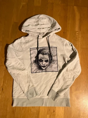 Limitato hoodie - Säljer en vit hoodie med stor huva och svart snörning. På bröstet finns ett stort tryck av Jokern i svart och grått, vilket ger en edgy och unik look. Hoodien har ribbade muddar vid ärmslut och nederkant. Perfekt för dig som gillar streetwear och grafiska motiv.