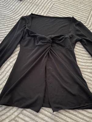 Svart långärmad topp med twist - Trendig svart långärmad topp med twistdetalj framtill och lätt v-ringad hals. Tunn och mjuk i materialet, perfekt för att styla med jeans eller kjol. Snygg croppad passform och enkel att matcha till olika looks.