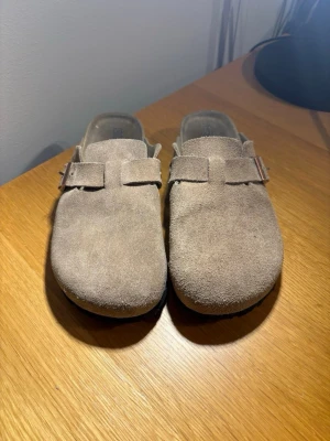 Birken stock - Säljer ett par snygga beige tofflor från birken stock med klassiskt spänne över foten, perfekta för en avslappnad stil hemma eller ute. Materialet är mjukt och ger en skön känsla. 