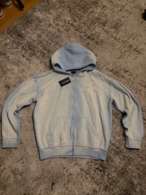 Lukamachain Hoodie - Ljusblå hoodie med dragkedja från Lukamachain. Hoodien har huva, fickor framtill och broderad text på bröstet samt stor broderad logga på ryggen. Mjuk bomullskänsla och ribbade muddar vid ärmar och midja. Perfekt för en chill och avslappnad stil. Pris kan diskuteras vid snabbt köp.