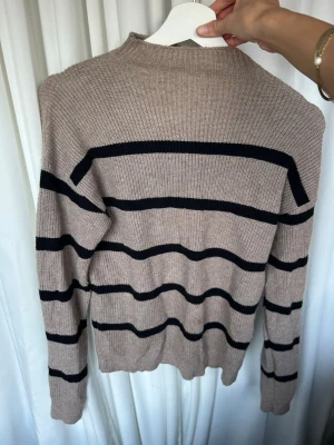 Beige stickad tröja med svarta ränder - Stickad tröja i beige med breda svarta horisontella ränder. Tröjan har hög hals och långa ärmar med ribbade muddar. Perfekt för lager på lager och enkel att matcha till olika outfits.