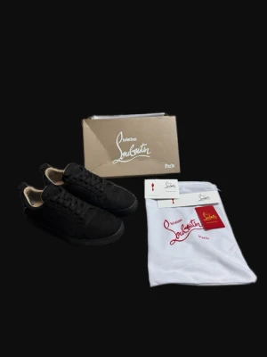 Svarta sneakers från Christian Louboutin - Snygga svarta sneakers från Christian Louboutin med klassisk röd sula och diskret logga. Skorna har en låg silhuett, svarta skosnören och ovandel i mocka. Perfekta för dig som vill ha en stilren och exklusiv look.
