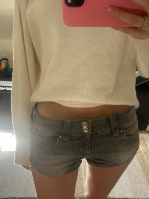 Grå lågmidjade jeansshorts - Säljer ett par grå denim minishorts med låg midja och dubbla knappar framtill. Shortsen har klassiska fem fickor och silver knappar. Perfekta för dig som gillar en edgy och trendig stil.