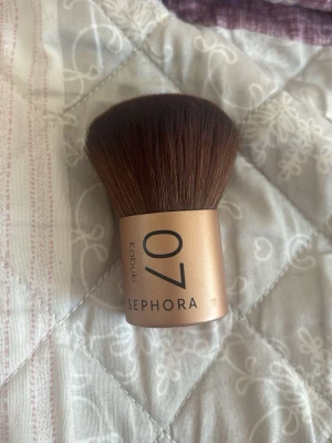 Kabukiborste Sephora 07 - Kabukiborste från Sephora, modell 07. Borsten har täta, mjuka strån i mörkbrunt och ett kompakt, rundat borsthuvud. Handtaget är kort och i en snygg guldig nyans med svart text. Perfekt för applicering av puder eller bronzer.