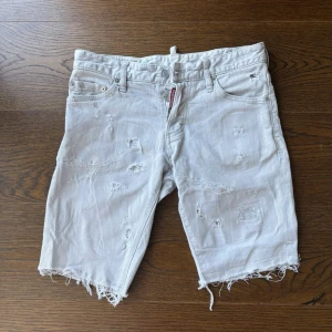 Vita distressed shorts från Dsquared2 - Säljer ett par vita shorts från Dsquared2 med slitna detaljer och råa kanter. Klassisk femficksmodell med knappgylf och Dsquared2-logga vid gylfen. Perfekta för en avslappnad och trendig look i sommar.