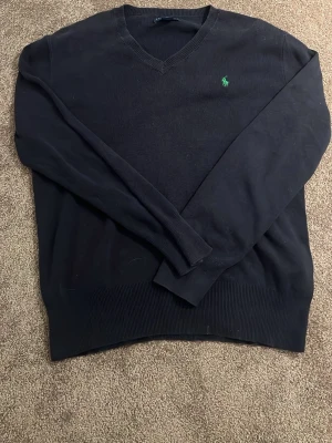 Svart stickad  tröja med från Polo Ralph Lauren - Klassisk svart tröja från Polo Ralph Lauren med V-ringning och den ikoniska gröna logon broderad på bröstet. Tröjan har långa ärmar och ribbade muddar vid ärmslut och nederkant. Perfekt för en stilren och enkel look.