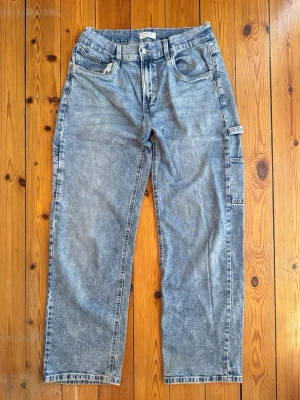 Blå barn cargobyxor i denim - Säljer ett par blå barn cargobyxor i tvättad denim med raka ben och flera praktiska fickor på sidorna och bak. Byxorna har bälteshällor och klassisk cargomodell med avslappnad passform. Perfekta för en streetwear-look.