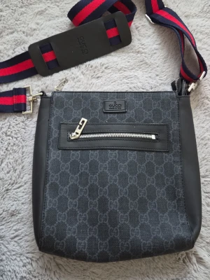 Axelväska   -  GG-mönster och detaljer i läder. Väskan har en framficka med silverfärgad dragkedja och justerbar axelrem i rött och blått. Perfekt för dig som vill ha en stilren och trendig accessoar.