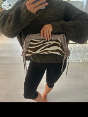 Kuvertväska med zebramönster - Snygg kuvertväska med zebramönstrad framsida i brunt och vitt. Väskan har beige virkade kanter och dekorativa snören på hörnen. Jag har gjort den själv så tyg och garn kostar en del + tiden att göra denna väska ❤️ 