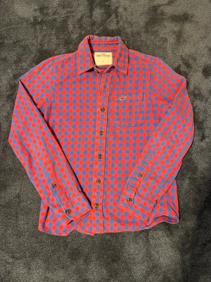 Vintage Hollister flannel röd/blå - Säljer en rutig skjorta från Hollister i rött och blått. Skjortan har klassisk krage, knappstängning framtill och en bröstficka med Hollister-loggan broderad. Långärmad och i mjuk bomull, perfekt för en avslappnad stil. Storlek: S, om det finns några frågor är det bara att skjuta ett meddelande. Fraktar snabbt!