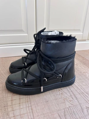 Svarta  boots från INUIKII ”Gloss” - Säljer ett par svarta moon boots från INUIKII med glansiga detaljer, grova snören och mjukt foder. Skorna har högre skaft och cool design med metallöglor och robust sula. Perfekta för kalla dagar när du vill hålla stilen. Köpta nya i butik av mig.