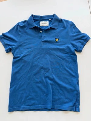 Blå pikétröja från Lyle & Scott - Säljer en klassisk blå pikétröja från Lyle & Scott med korta ärmar och gul broderad logga på bröstet. Tröjan har krage och knappar framtill, perfekt för en avslappnad men ändå snygg stil.