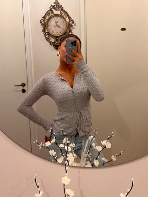 Ljusgrå kabelstickad kofta - Supersöt ljusgrå kabelstickad kofta med knytband i midjan och v-ringad hals. Koftan har långa ärmar och ett diskret mönster som ger en trendig look. Perfekt att matcha med jeans eller kjol för en chill och snygg outfit.