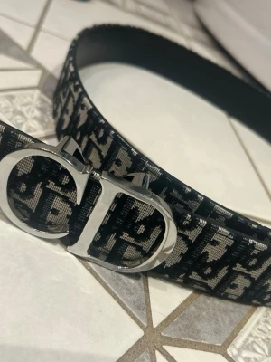 Svart mönstrat bälte med silverdetalj - Snyggt svart bälte med ett grafiskt mönster i beige och stor silverfärgad spänne med bokstäver. Perfekt accessoar för att lyfta vilken outfit som helst. Bältet har en modern och trendig vibe.