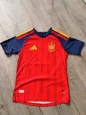 Spanien fotbollströja  - ÄKTA FOTBOLLSTRÖJA, Säljer en officiell matchtröja för Spaniens landslag från Adidas. Tröjan är röd med marinblå ärmar och gula detaljer på axlarna. Den har ett tryckt landslagsemblem och Adidas-logga på bröstet. Tillverkad i lätt och ventilerande funktionsmaterial.